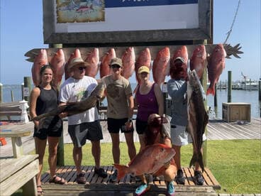 A group of anglers proudly displaying their catch, including a large Grouper and several Snapper, in front of a fishing charter sign on the Gulf Coast.