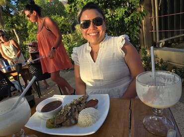 A group of friends enjoying a meal outdoors, with one person smiling at the camera while seated at a table.