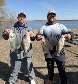 Two anglers proudly displaying their catch of Crappie by the lakeside, showcasing a successful day of fishing.