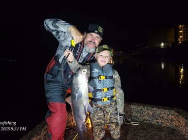 A proud father and son duo displaying a large catfish during a nighttime fishing adventure.