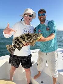 Two anglers proudly displaying a large Goliath Grouper while fishing in clear waters, showcasing a successful day on the boat.