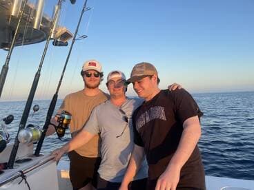 Three friends enjoying a fishing trip on a boat, with fishing rods visible in the background and a scenic ocean view.