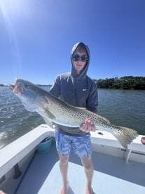 Young angler proudly holding a large Speckled Trout while fishing on a boat in clear waters.
