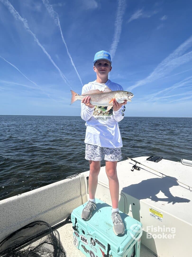 Young angler proudly holding a Redfish while standing on a cooler in a boat, enjoying a sunny day on the water.