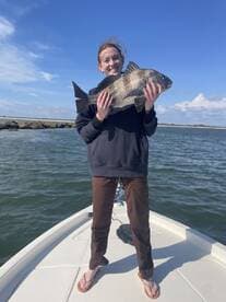 A young angler proudly displaying a large Black Drum while standing on the bow of a boat, enjoying a sunny day on the water.