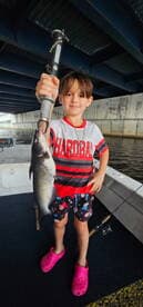 A young angler proudly holding a sizable catfish while fishing under a bridge, showcasing a fun day on the water.