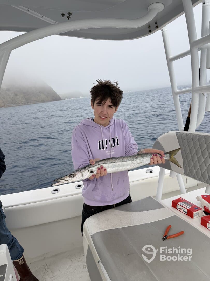 A young angler proudly displaying a Barracuda while fishing on a boat in foggy conditions.