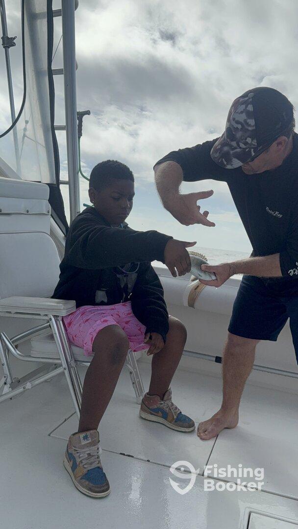 A young angler is receiving guidance on handling a fish while seated on a boat, showcasing a hands-on fishing experience.