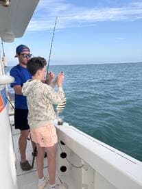 A young angler and an adult are actively fishing on a boat, enjoying a day on the water with a beautiful ocean backdrop.