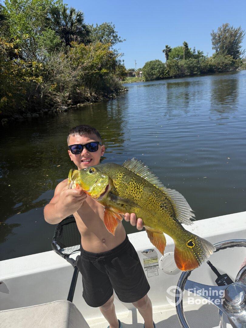 A young angler proudly displaying a vibrant Peacock Bass while fishing on a sunny day in a scenic waterway.