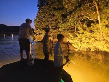 Three anglers, including a child, are fishing at dusk from a boat, illuminated by the glow of the water and surrounding trees.