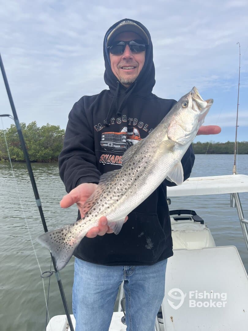 Angler proudly displaying a Speckled Trout while fishing from a boat in a scenic waterway.