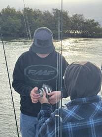 Two young anglers on a fishing trip, one proudly holding a striped fish while the other assists, surrounded by a scenic waterway.