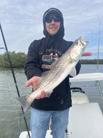 Angler proudly displaying a Speckled Trout while fishing from a boat in a scenic waterway.