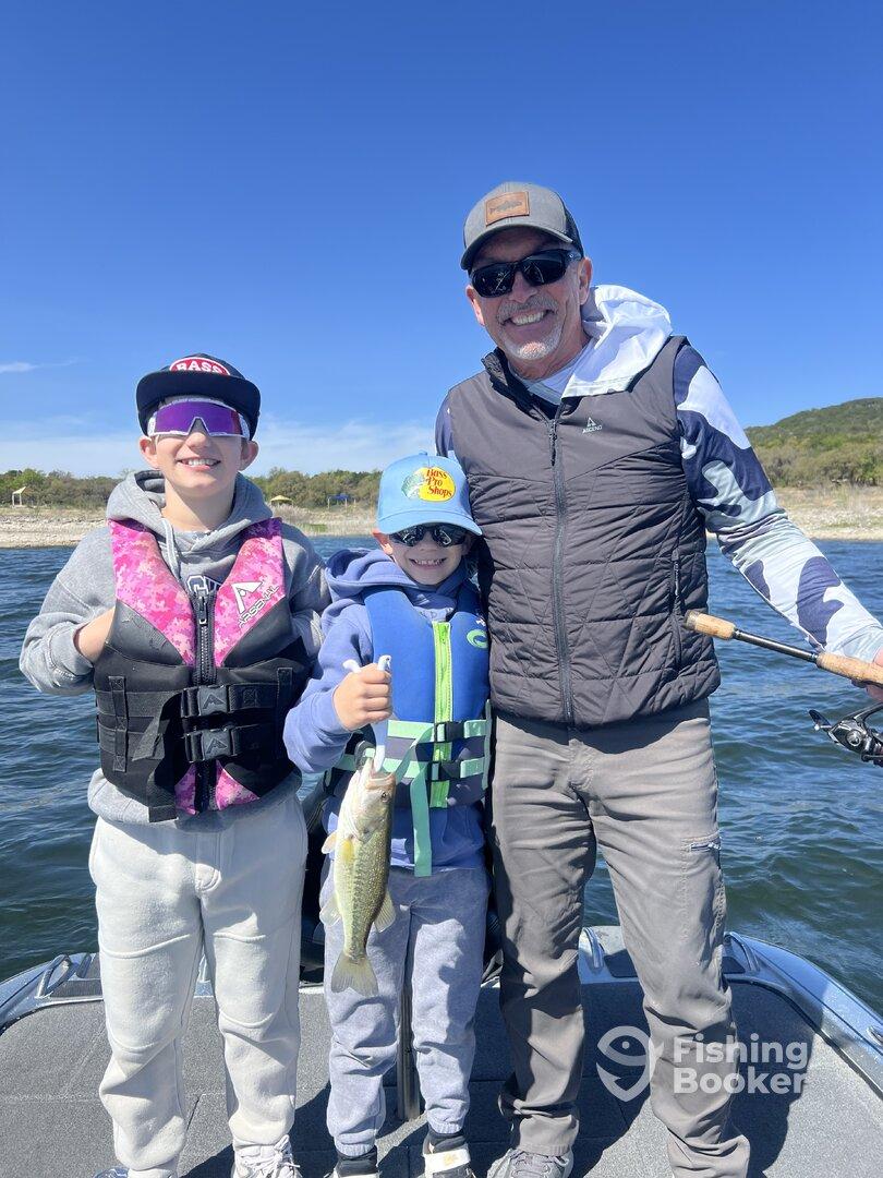 A family enjoys a successful fishing trip, showcasing a young angler holding a Largemouth Bass while posing with a sibling and an adult on a boat.