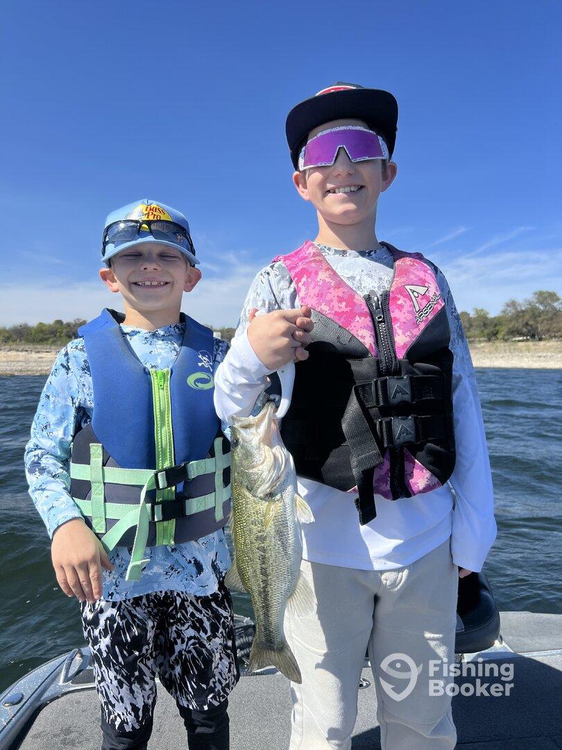Two young anglers proudly displaying a large Largemouth Bass while fishing on a sunny day.