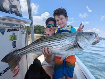 A young boy proudly holds a large striped bass while posing with an adult on a boat, showcasing a fun family fishing experience.