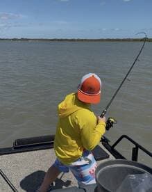 A young angler in a bright yellow hoodie and colorful shorts is reeling in a fish from a boat in a calm waterway, showcasing the excitement of fishing.