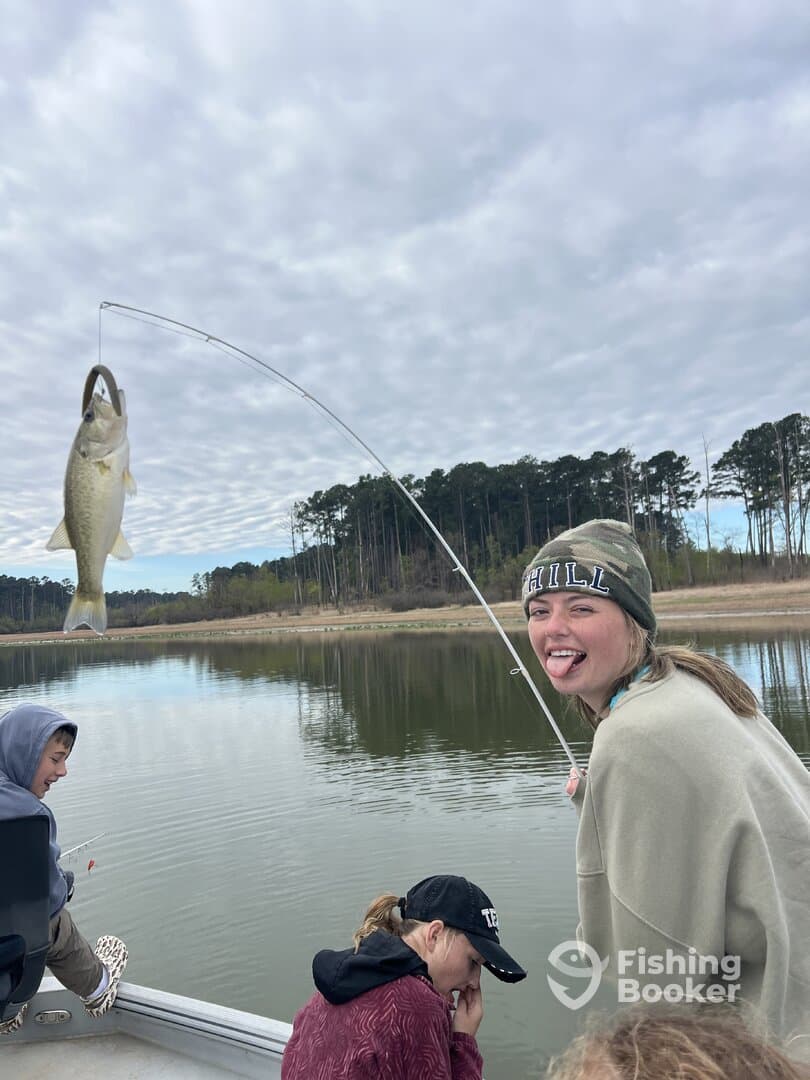A group of young anglers enjoying a day of fishing on a calm lake, with one girl playfully reeling in a large bass.