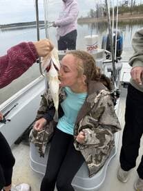 A young angler joyfully kisses a freshly caught fish while seated on a boat, surrounded by friends on a fishing trip.