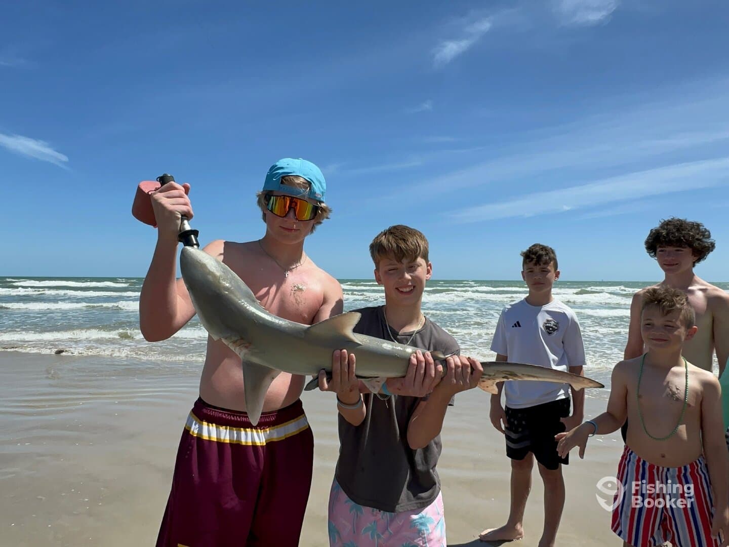 A group of young anglers proudly displaying a caught shark on the beach, showcasing their successful fishing trip.