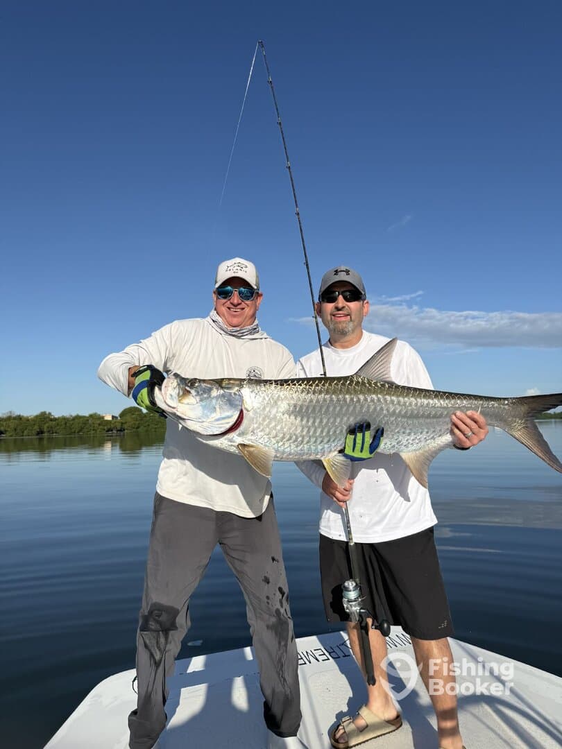 Two anglers proudly displaying a large Tarpon while fishing on a calm day in a scenic waterway.
