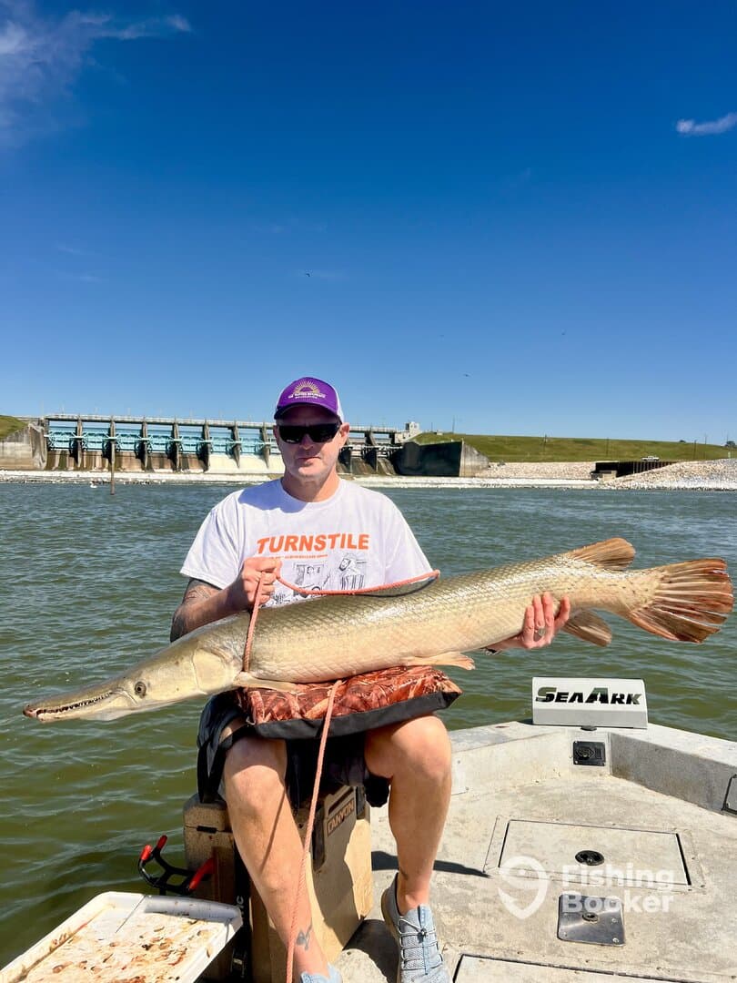 Angler proudly displaying a large Northern Pike while seated in a SeaArk boat, with a dam visible in the background.