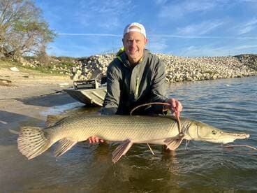 Angler proudly displaying a large Northern Pike caught during a fishing trip, with a scenic backdrop of a rocky shoreline.