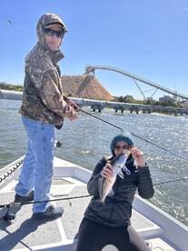 Angler proudly displaying a Speckled Trout while fishing on a boat, with a scenic industrial backdrop.