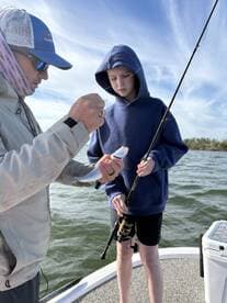 A young angler watches closely as an adult prepares to unhook a freshly caught Speckled Trout while fishing on a boat.