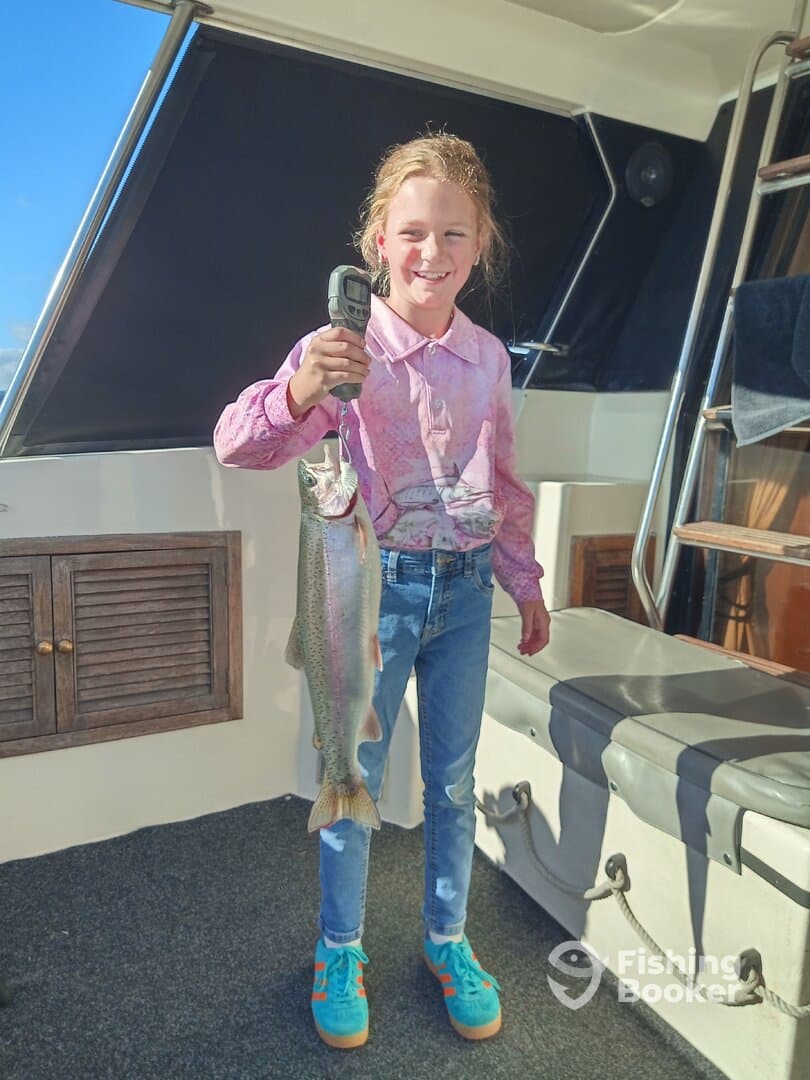A young angler proudly displaying a large Rainbow Trout aboard a fishing boat, showcasing a successful catch during a family fishing trip.