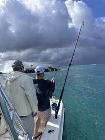 Two anglers actively fishing from a boat, with rods bent as they reel in their catch near rocky outcroppings in clear blue waters.