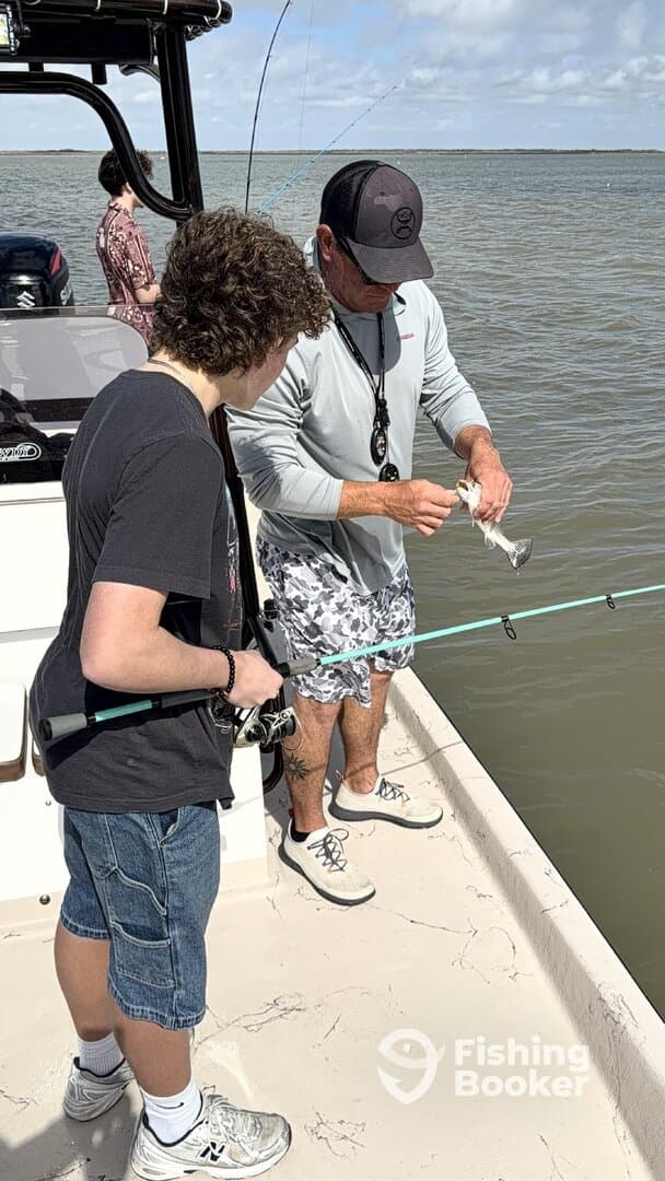 A young angler and an adult are engaged in fishing on a boat, with the adult demonstrating how to handle a small fish caught in the water.