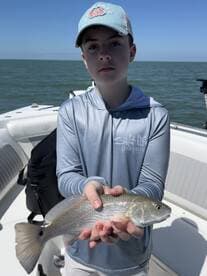 A young angler proudly displaying a caught Redfish while on a boat in a sunny fishing environment.
