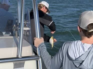 A young angler is excitedly reeling in a fish while another person assists on a boat, showcasing a lively fishing experience.