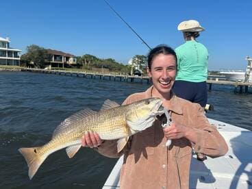 A young woman proudly displays a large Redfish while fishing from a boat, with a scenic waterfront in the background.