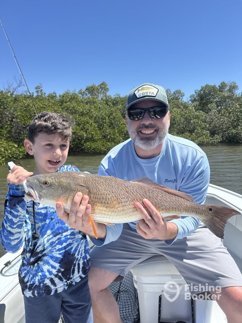 A young angler and an adult proudly display a Redfish caught during a fishing trip in a scenic coastal environment.
