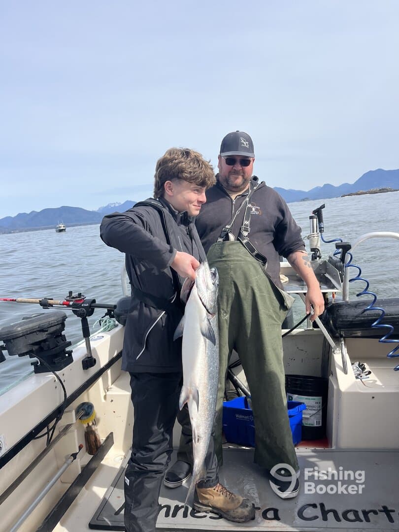 A young angler proudly displaying a large fish alongside an adult on a fishing charter in a scenic coastal environment.