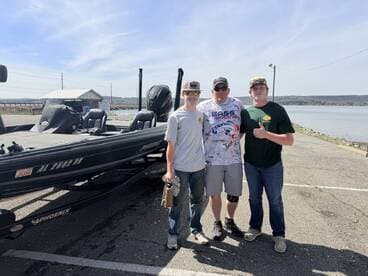 Three anglers posing next to a Phoenix bass boat at a lakeside launch, showcasing a sunny day and a friendly atmosphere.