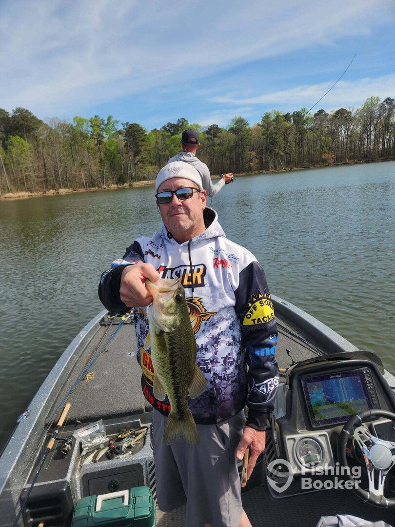 Angler proudly displaying a Largemouth Bass while fishing on a serene lake, with another fisherman casting in the background.