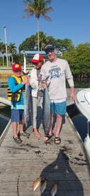 A family proudly displaying their catch of two large fish at the dock, enjoying a sunny day in a tropical fishing environment.