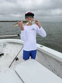 Young angler proudly holding a Speckled Trout while fishing from a boat in a coastal environment.
