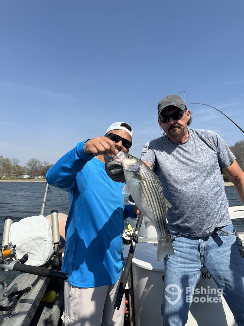 Two anglers proudly displaying a large Striped Bass while fishing on a sunny day.