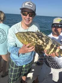 A young angler proudly displaying a large Grouper while fishing with family on a sunny day.