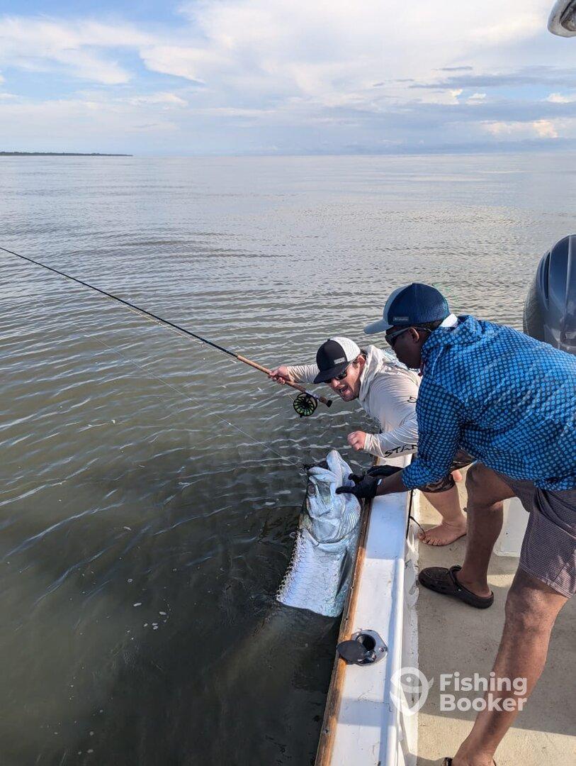 Two anglers working together to reel in a large fish while fishing in calm waters, showcasing an exciting moment on the boat.