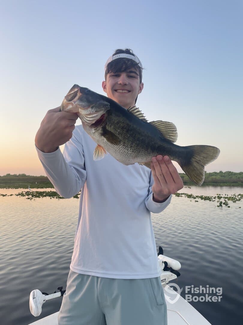 A young angler proudly displaying a large Largemouth Bass while fishing in a serene lake setting at sunrise.