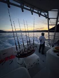 A fisherman stands on the deck of a sportfishing boat, surrounded by fishing rods, as the sun sets over the water.