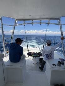 Two anglers seated in the helm area of a sportfishing boat, preparing for a day of deep-sea fishing in clear blue waters.
