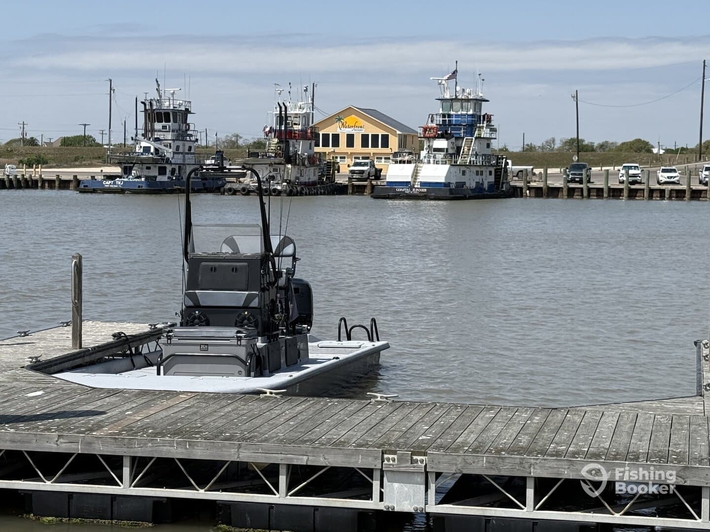 A sleek fishing boat docked at a marina, with tugboats and a waterfront establishment in the background.