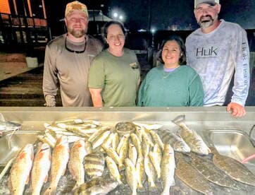 A group of anglers proudly displaying their catch of various fish, including Redfish and Flounder, arranged on a cleaning table after a successful night fishing trip.
